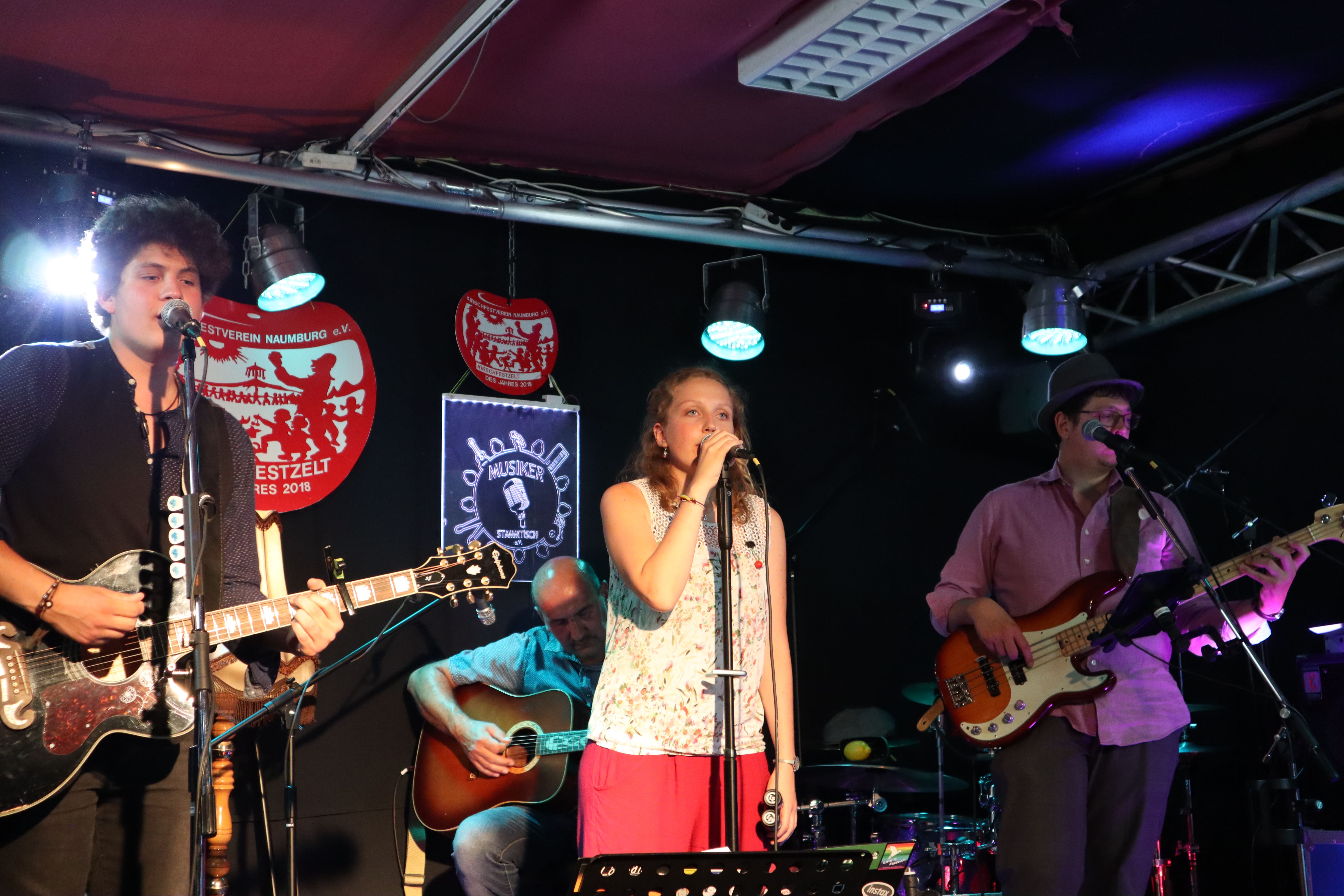 Auf dem Bild ist die Band bei dem Auftritt zum Kirschfest 2020 zu sehen. Charlotte singt. Tobi singt und spielt Gitarre. Henrik spielt, im Hintergrund, auch Gitarre. Christoph spielt, ganz rechts außen, Bass.